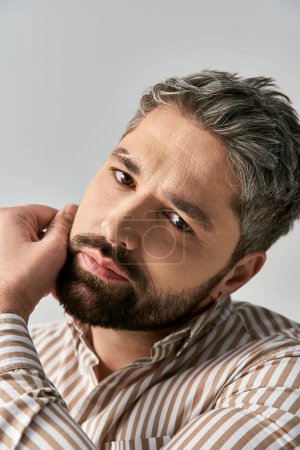 A stylish man with a beard exuding charm in a striped shirt against a neutral studio backdrop.