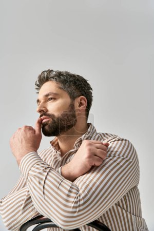 Photo for A sophisticated man with a beard poses confidently in a striped shirt against a grey studio backdrop. - Royalty Free Image