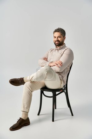Téléchargez les photos : Un homme barbu en tenue élégante est assis sur une chaise avec les jambes croisées dans un studio sur un fond gris. - en image libre de droit