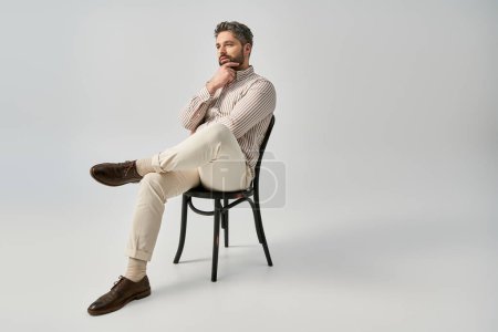 A stylish man with a beard and elegant attire sits on top of a chair with confidence against a grey studio background.