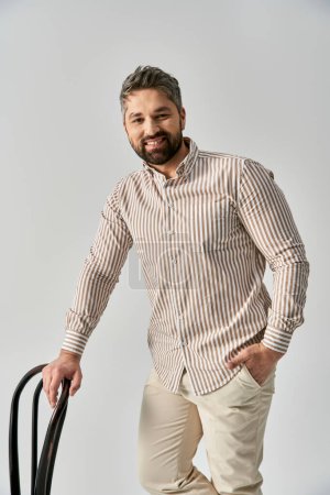 Foto de Un hombre elegante con una barba de pie con confianza, sosteniendo una maleta, y sonriendo con alegría en un fondo gris estudio. - Imagen libre de derechos