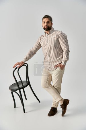 A bearded man in elegant attire striking a pose next to a sleek black chair against a grey studio background.