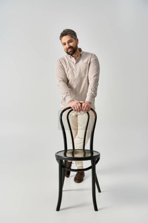 Foto de Un hombre elegante con barba en un elegante atuendo cerca de una elegante silla negra sobre un fondo gris neutro. - Imagen libre de derechos
