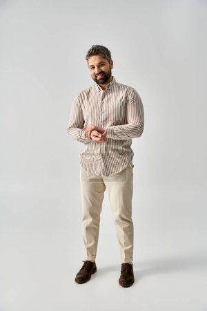 A stylish man with a beard dressed in elegant attire stands confidently against a grey studio background.