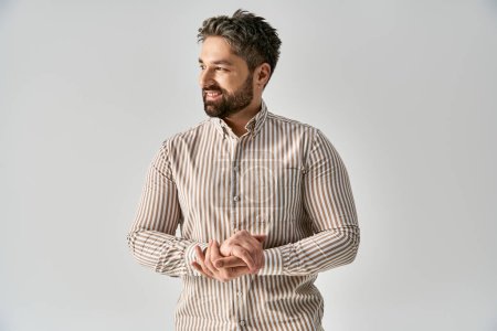 Photo for A bearded man stands with folded hands, exuding elegance and poise in fashionable attire against a grey studio backdrop. - Royalty Free Image