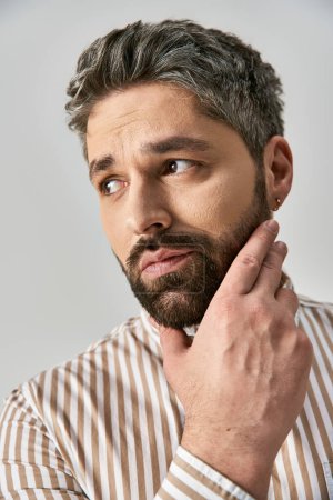 A captivating man with a beard poses in elegant attire against a grey background in a studio setting.