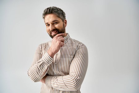 A stylish man with a beard poses in elegant attire against a grey background in a studio setting.