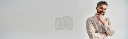 Photo for A stylish man with a beard and elegant attire stands with hands on chin, showcasing his alluring appearance on a grey background. - Royalty Free Image