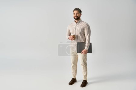 Téléchargez les photos : Un homme barbu en tenue élégante tenant un ordinateur portable et une tasse de café, mettant en valeur un équilibre entre travail et détente. - en image libre de droit