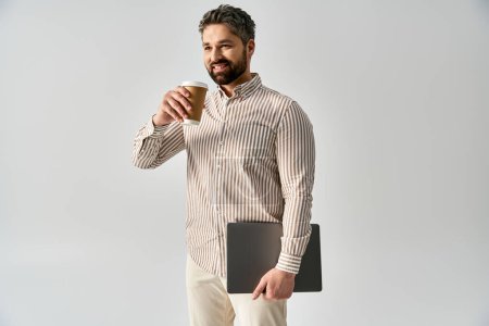 A stylish man in elegant attire holding a laptop and a cup of coffee against a grey background.