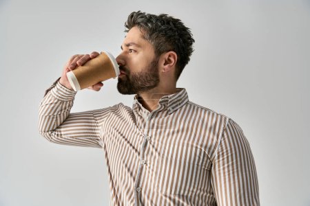 A dapper man with a beard enjoying a drink from a paper cup against a grey backdrop in a studio setting.