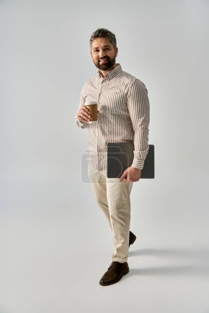 A bearded man in elegant attire holding a laptop and a cup of coffee, exuding sophistication and relaxation.