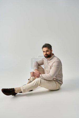 Téléchargez les photos : Un homme barbu en tenue élégante assis sur le sol les jambes croisées dans une pose contemplative sur un fond de studio gris. - en image libre de droit