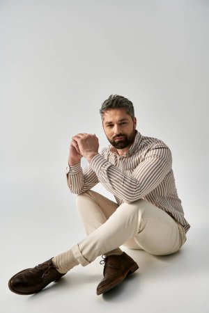Ein bärtiger Mann in eleganter Kleidung sitzt im Schneidersitz auf dem Boden in einem Studio vor grauem Hintergrund und strahlt Gelassenheit aus..
