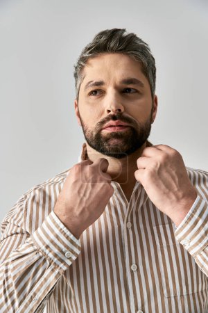 Un hombre barbudo emana confianza en una elegante camisa a rayas sobre un fondo gris de estudio.