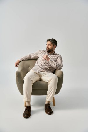 A stylish man with a beard sits in a chair, deep in thought with his hand on his chin, against a grey studio background.
