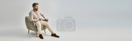 Foto de Un hombre elegante con barba y elegante atuendo se sienta en sillón sobre un fondo gris. - Imagen libre de derechos
