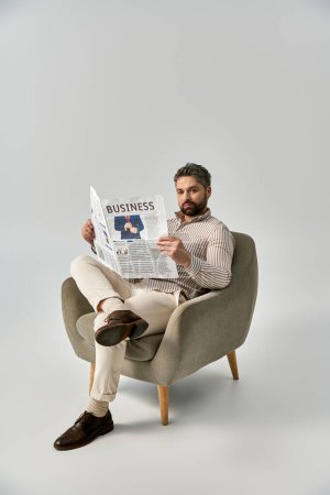 A stylish, bearded man in elegant attire sits in a chair reading a newspaper against a grey studio background.