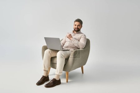 Téléchargez les photos : Un homme barbu en tenue élégante s'assoit dans une chaise, concentré sur son ordinateur portable. - en image libre de droit