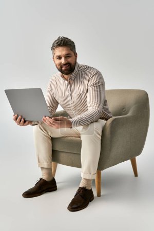 A stylish man with a beard is seated on a chair, working on his laptop in a trendy and sophisticated setting.