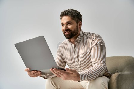 Téléchargez les photos : Un homme élégant avec une barbe assise sur une chaise tout en tenant un ordinateur portable dans un cadre de studio sur un fond gris. - en image libre de droit