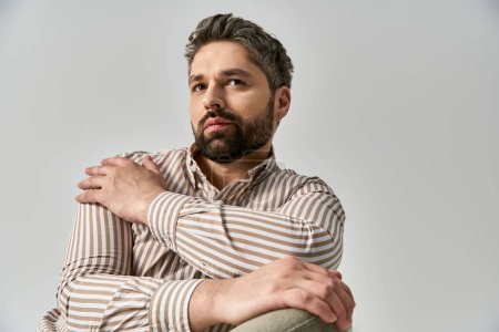 Un hombre elegante con barba posando con una camisa a rayas sobre un fondo gris de estudio.