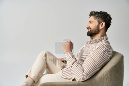 Ein stilvoller bärtiger Mann in eleganter Kleidung sitzt auf einem Sessel vor grauem Studiohintergrund.