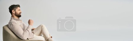 Photo for A fashionable man with a beard sits in armchair, crossing his legs, wearing elegant attire, captured against a grey studio backdrop. - Royalty Free Image