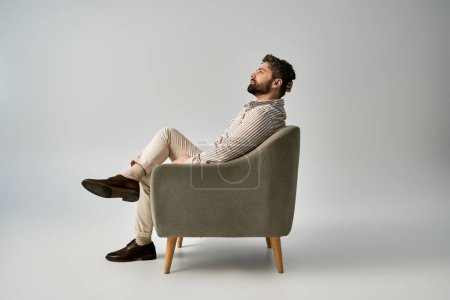 A stylish man with a beard sits in a chair, crossing his legs, showcasing elegance and confidence.