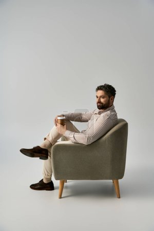 A dapper man with a beard relaxes in a chair, savoring a cup of coffee in an elegant fashion.