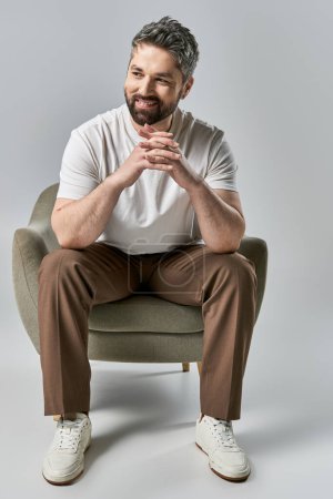 Un hombre elegante con barba se sienta en una silla con las manos dobladas, exudando una sensación de tranquilidad y pensamiento profundo.