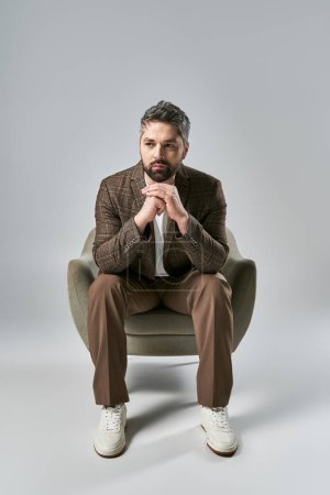 Foto de Un hombre con barba sentado en una silla, las manos dobladas en una pose elegante sobre un fondo gris de estudio. - Imagen libre de derechos
