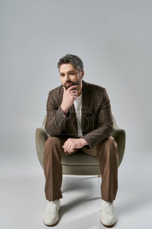 A bearded man in elegant attire sits with hand on chin, pondering deeply in a stylish pose against a grey studio background.