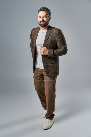 A captivating man with a beard strikes a pose in an elegant brown suit and white shirt against a grey studio backdrop.
