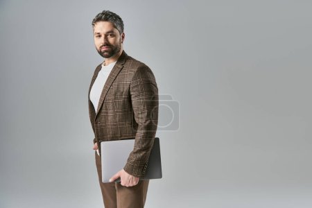 Téléchargez les photos : Homme élégant en costume et cravate tenant avec confiance un ordinateur portable sur un fond de studio gris. - en image libre de droit