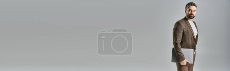 Téléchargez les photos : Un homme barbu en costume se tient debout avec confiance, tenant un ordinateur portable, respirant élégance et style sur un fond gris. - en image libre de droit