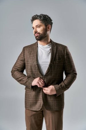A charismatic man with a beard wearing a brown jacket and pants strikes a pose in an elegant fashion on a grey studio backdrop.