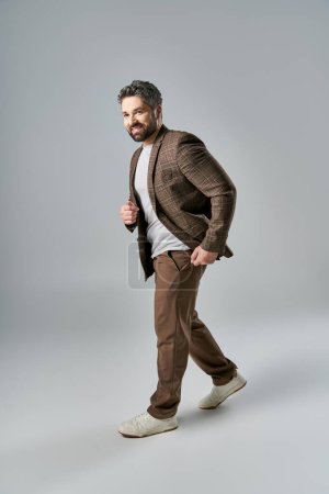A bearded man strikes a pose in an elegant brown jacket and crisp white shirt against a grey background in a studio setting.