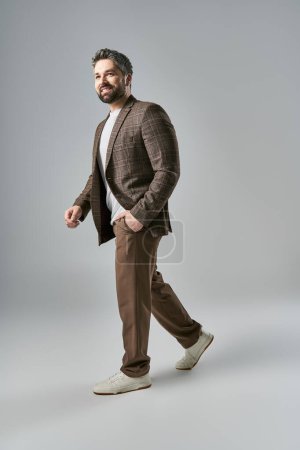 A stylish man with a beard in an elegant brown suit and white shirt striking a pose against a grey studio backdrop.