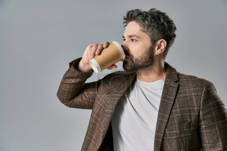 Foto de Un hombre carismático con barba sosteniendo elegantemente una taza de café en la mano, exudando sofisticación sobre un fondo gris de estudio. - Imagen libre de derechos