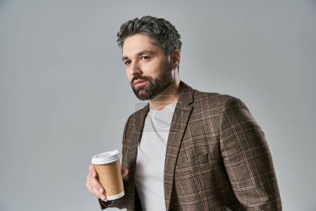 Téléchargez les photos : Un homme élégant avec une barbe tient élégamment une tasse de café, exsudant charme et sophistication sur un fond gris. - en image libre de droit
