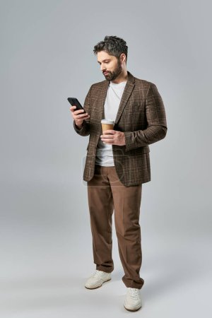 A stylish man in elegant attire holding a coffee cup and checking his phone while lost in thought.