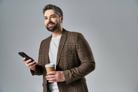 Foto de Un hombre barbudo con elegante atuendo sostiene una taza de café mientras revisa su teléfono celular, exudando sofisticación y multitarea. - Imagen libre de derechos