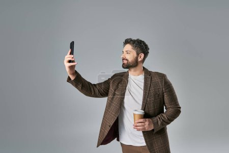 A stylish man with a beard dressed in elegant attire taking a self-portrait with his cell phone against a grey background.