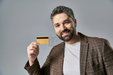 A stylish man with a beard holding a credit card and smiling joyfully against a grey studio background.