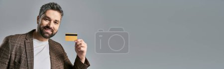 Téléchargez les photos : Un homme élégant avec une barbe tenant une carte de crédit sur fond de studio gris. - en image libre de droit