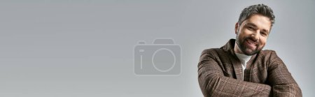 Ein bärtiger Mann in brauner Jacke posiert selbstbewusst in einem Studio vor grauem Hintergrund.