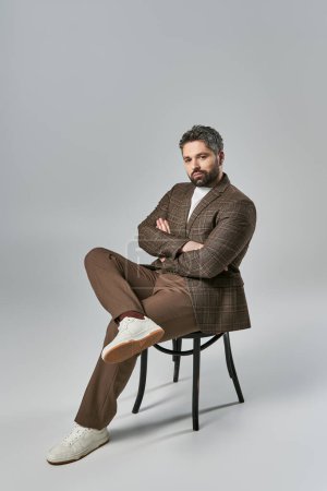 Un homme barbu est assis avec les bras croisés, respirant la force et l'équilibre dans une tenue élégante sur fond de studio gris.