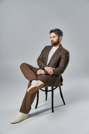 A sophisticated man with a beard sits elegantly on a chair, crossing his legs, in a studio with a grey background.