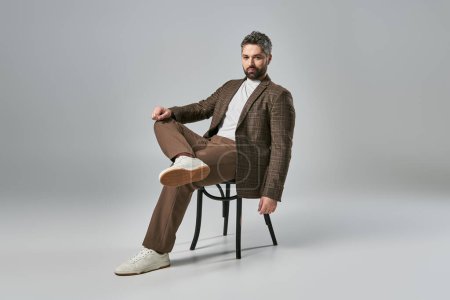Téléchargez les photos : Un homme élégant avec une barbe est assis sur une chaise, croisant ses jambes élégamment dans une pose avant-gardiste sur un fond de studio gris. - en image libre de droit